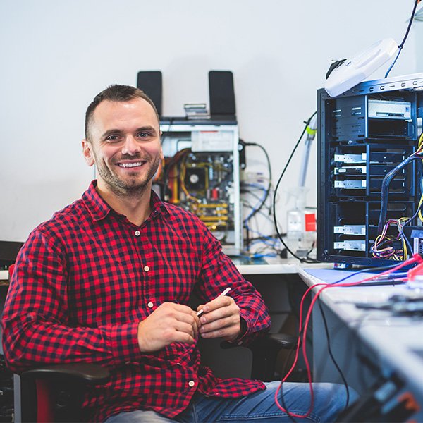 Hombre de camisa a cuadros rojos y negros sonriendo mientras está sentado en un escritorio. A su lado hay una computadora abierta con cables visibles y componentes electrónicos expuestos. El fondo muestra otra computadora parcialmente ensamblada.